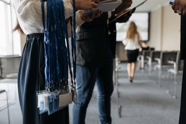 Une photo de deux organisateurs d'un salon professionnel, centrée sur les badges bleus des participants pendant au bras de l’une d’eux, et en arrière-plan, une femme de dos se dirigeant vers son siège pour assister à une ou plusieurs conférences IA.